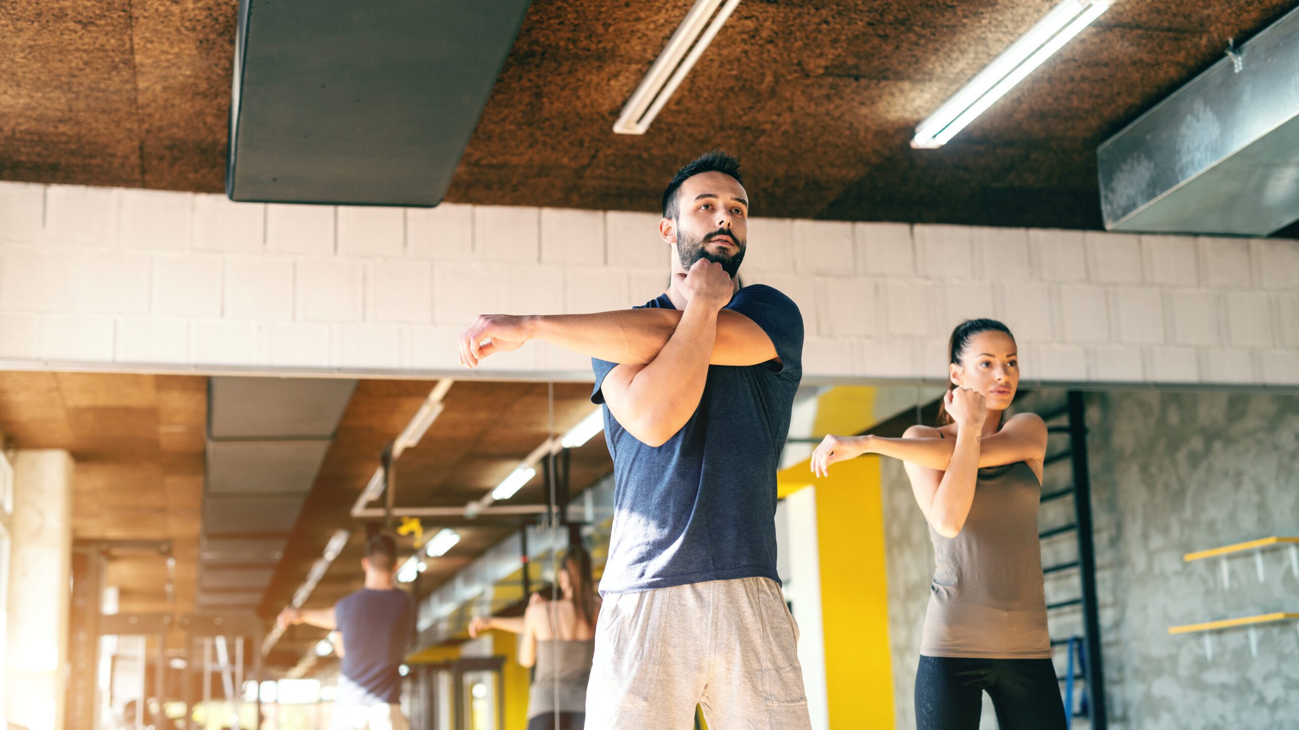 Cute young sporty Caucasian couple in sportswear stretching arms in gym. In background mirror.